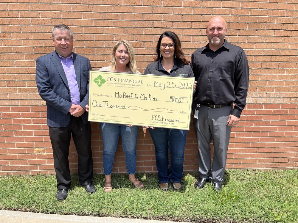 Male in suit, 2 females holding a check, male in gray shirt and pants standing in front of a brick wall.
