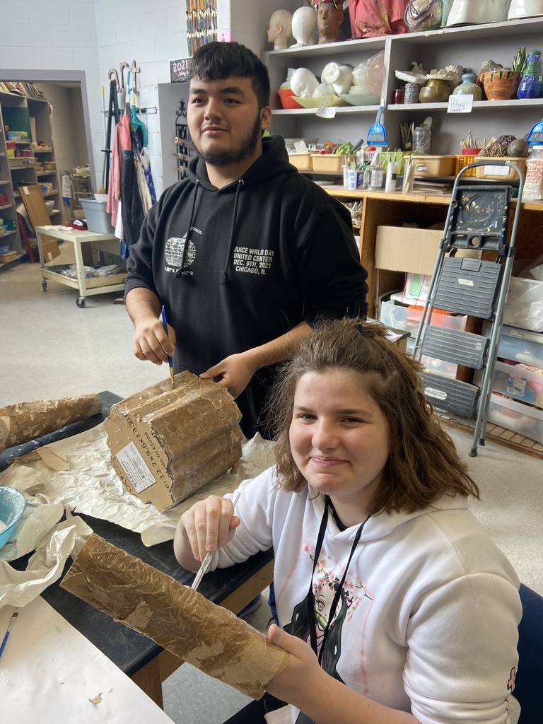 Female student wearing white hoodie, male student wearing black hoodie, work together on their sculpture art projects. 